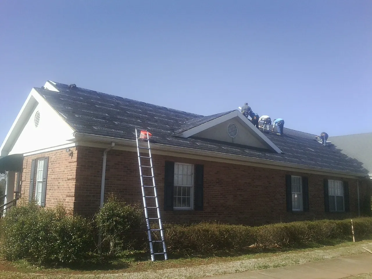 Crew tearing shingles off a brick ranch roof with a ladder set — Metal Roof Repair work by our crew in University